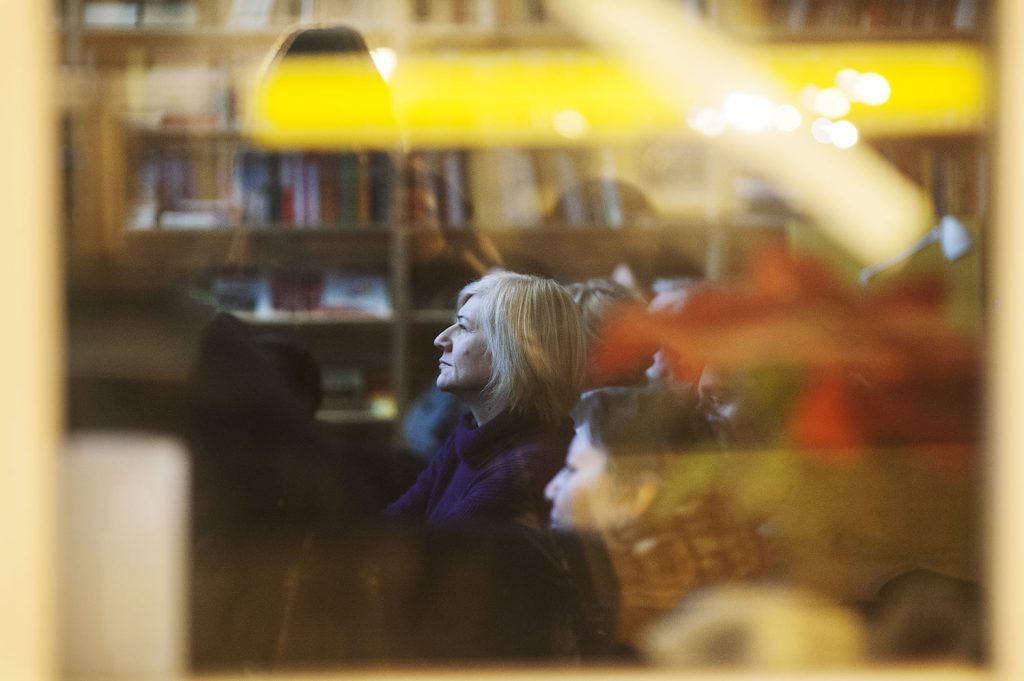 Kobieta o blond włosach, siedząca wśród innych, uważnie słucha w słabo oświetlonym pokoju z półkami na książki w tle, widzianym przez okno z odbiciami i rozmytymi światłami.