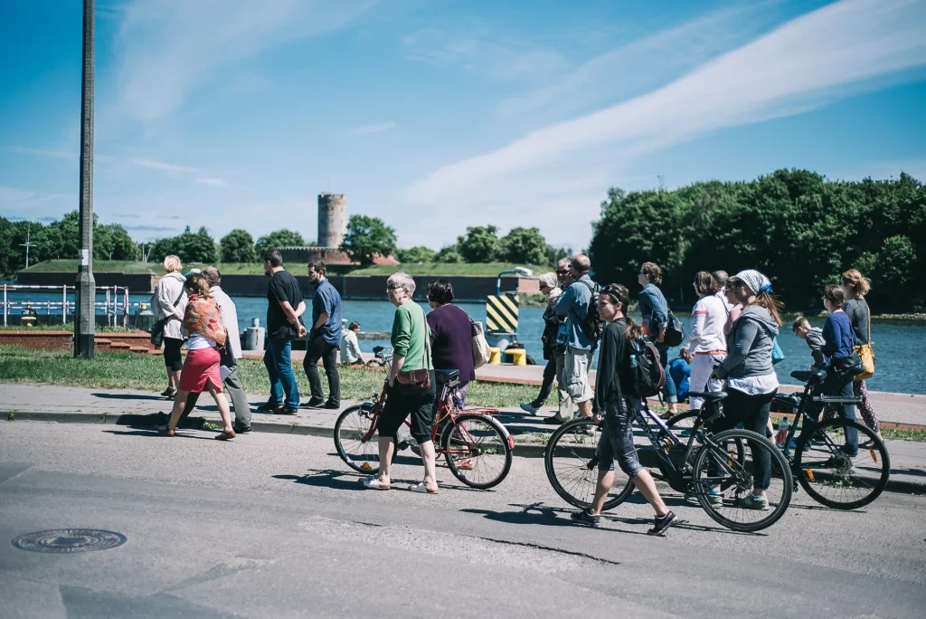 Grupa ludzi, niektórzy idą pieszo, a niektórzy na rowerach, przechodzą przez ulicę w pobliżu rzeki w słoneczny dzień. Drzewa i zabytkowa wieża są widoczne w tle pod błękitnym niebem z delikatnymi chmurami.