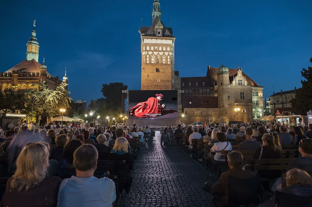 Duży tłum ogląda nocny seans filmowy na świeżym powietrzu, siedząc na ławkach na historycznym placu miejskim z oświetlonymi budynkami i wysoką wieżą zegarową w tle.