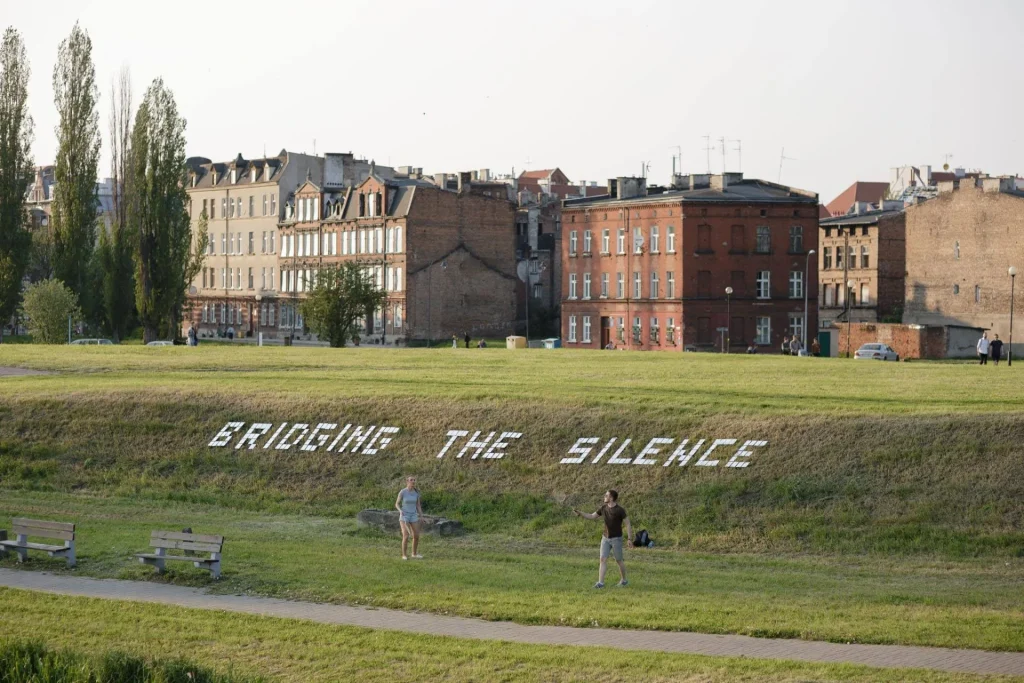 Trawiaste pole z dużymi białymi literami BRIDGING THE SILENCE stoi przed rzędem starych ceglanych budynków; dwie osoby stoją i chodzą w pobliżu, a ławki siedzą wzdłuż ścieżki na pierwszym planie.