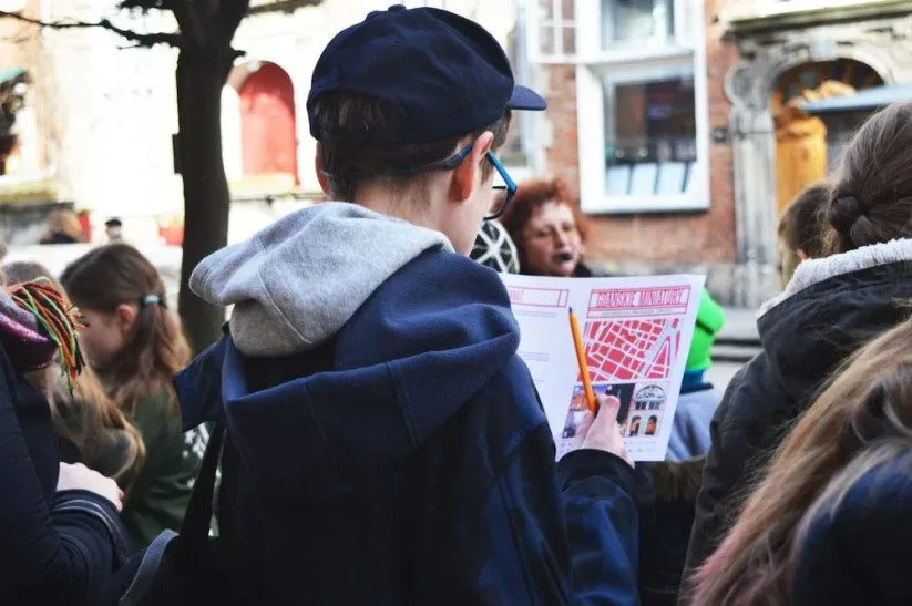 Osoba w granatowej czapce i kurtce trzyma mapę miasta, stojąc w grupie na zewnątrz, prawdopodobnie podczas pieszej wycieczki z przewodnikiem po obszarze miejskim. W tle widoczni są ludzie i zabytkowe budynki.
