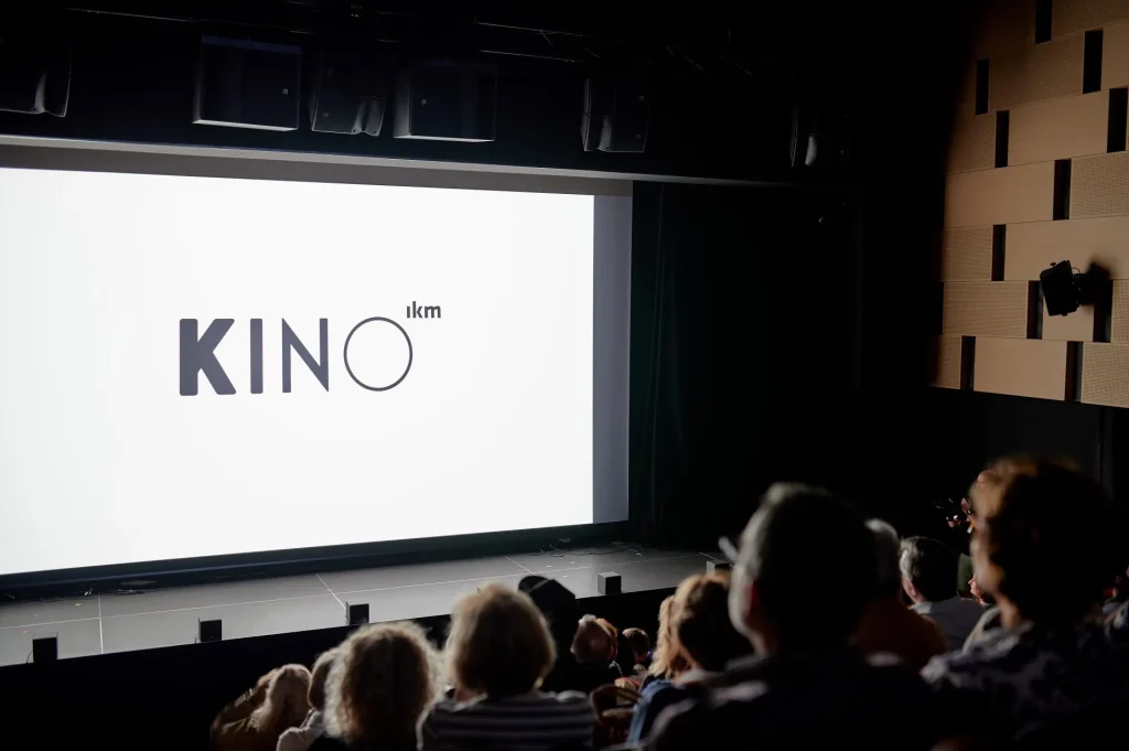 Grupa ludzi siedzi w ciemnej sali kinowej, oglądając duży ekran, na którym wyświetlane jest słowo KINO pogrubionymi literami na białym tle. Ściany kina mają nowoczesny, geometryczny design.