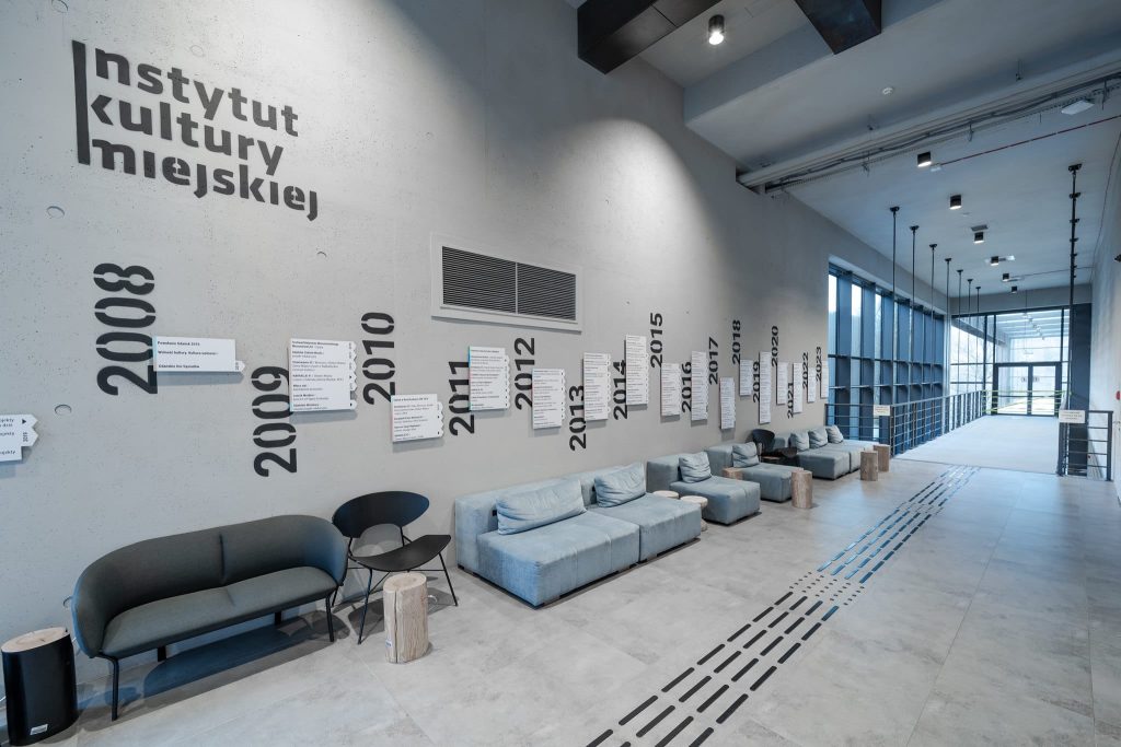 Nowoczesne lobby z jasnoszarymi ścianami i sofami, oznaczone dużymi czarnymi latami od 2008 do 2020 roku. Plakaty i napis Instytut Kultury Miejskiej w języku polskim podkreślają ikm praca Gdańsk. W korytarzu znajdują się duże okna.